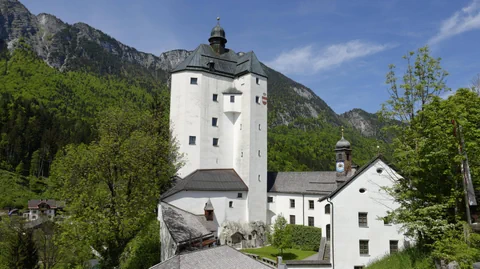 weisser Kirchenturm mit grauem Dach, davor weisses, dreistöckiges Gebäude. Dahinter Berge.