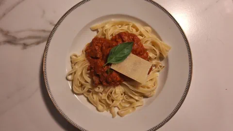 Ein weisser Teller mit Nudeln und roter Tomatensauce. Darauf liegt ein Basilikumblatt und ein Stück Parmesan. 