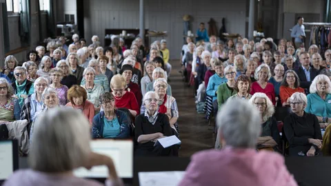 Eine grosse Gruppe älterer Menschen sitzt in einem Saal und hört einer Präsentation zu.