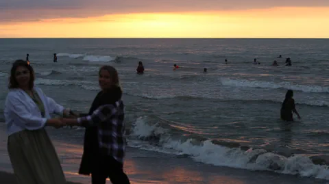 Zwei Frauen ohne Kopftuch tanzen am Strand, die Sonne geht unter.