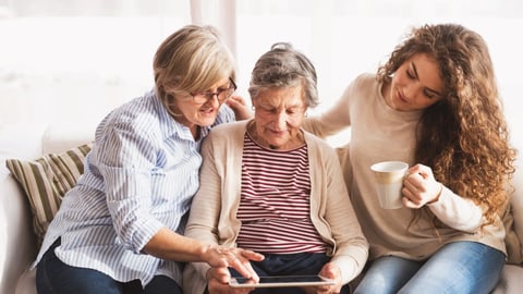 Frauen aus drei Generationen schauen auf ein Touchpad.