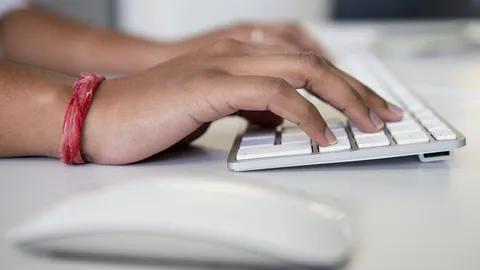 Hände, die auf einer Tastatur etwas eintippen. Die rechte Hand trägt ein rotes Band.