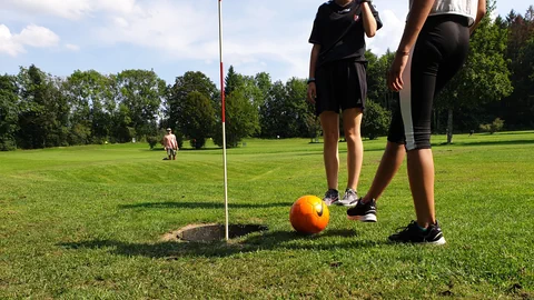 Zwei Personen beim Footgolf auf einer Wiese nahe eines Golflochs, Mann im Hintergrund.