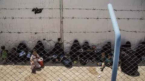 An einer Hauswand sitzen im Schatten Frauen und Kinder, davor ein Zaun mit Stacheldraht.