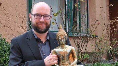 Mann mit Hemd, Veston, Brille, Bart und Buddha-Statur in der Hand