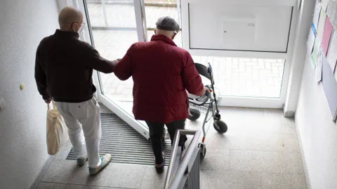 Person unterstützt ältere Person mit Rollator beim Verlassen eines Gebäudes.