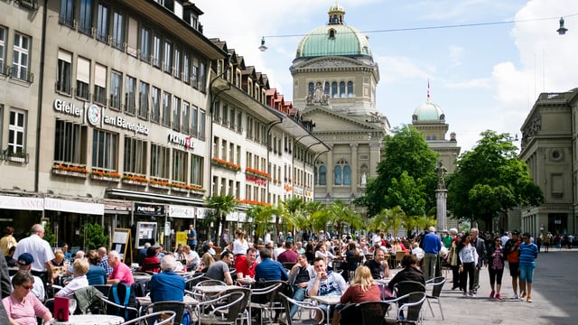 Menschen sitzen in Bern auf dem Bärenplatz in Strassenrestaurants, dahinter das Bundeshaus.