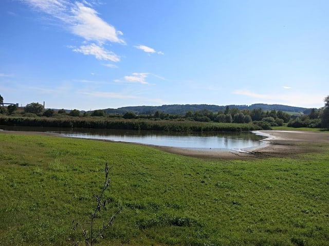 Landschaft mit Wiese, kleinem See und Hügeln im Hintergrund.