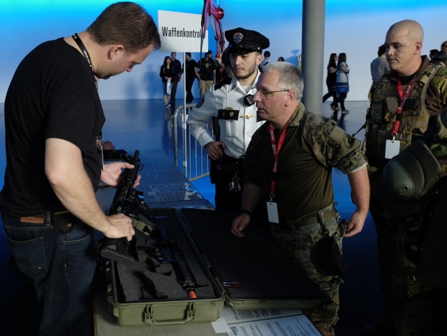 Männer in Uniform, einer kontrolliert ein Gewehr. Verkleideter Politist im Hintergrund.