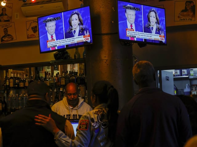 Menschen sehen sich in einer Bar eine Nachrichtensendung auf TV-Bildschirmen an.