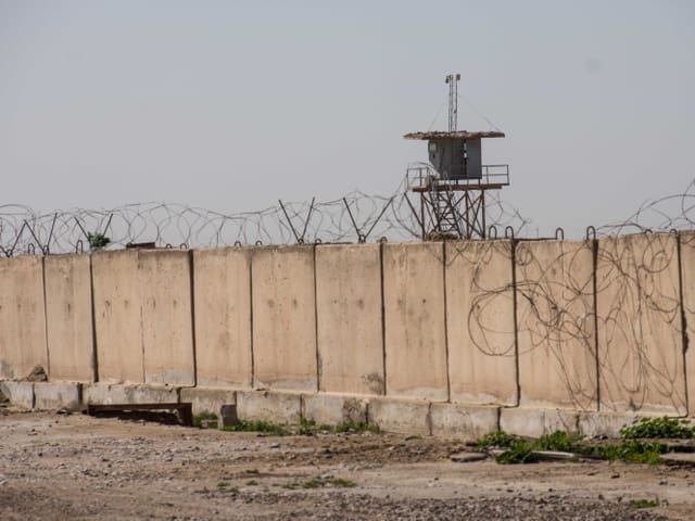 Eine Betonmauer gesäumt von Stacheldraht in der Wüste. Dahinter ein Wachturm.