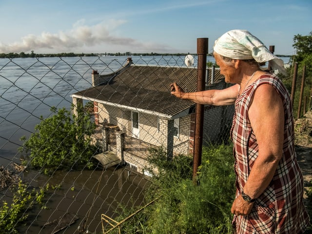 Eine alte Frau blick von einem kleinen Hügel auf den Fluss und ein Haus, das überschwemmt am Rand steht.