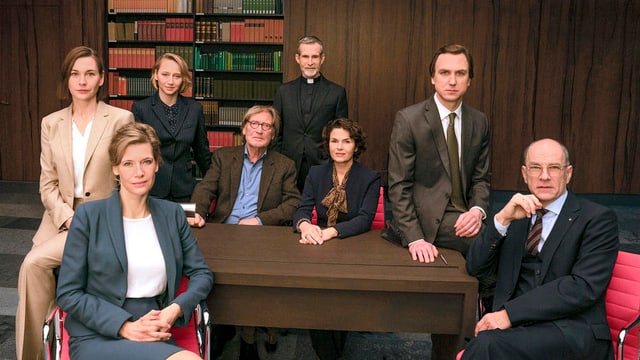 Eight men and women sit around a table and look into the camera.  They wear office clothes.