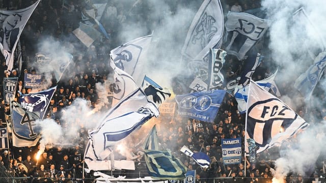 Fussballfans mit Fahnen und Rauch in Stadionkurve.