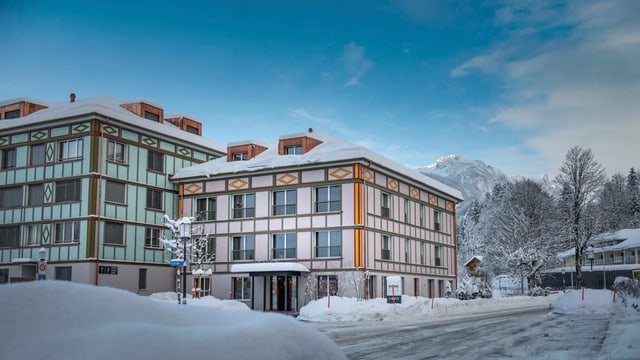 Ein eingeschneites Hotelgebäude. Im Hintergrund kahle, beschneite Bäume und ein Berggipfel. 