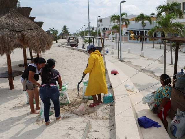 Menschen packen Sandsäcke an einem Strand mit Palmen und Hütten.