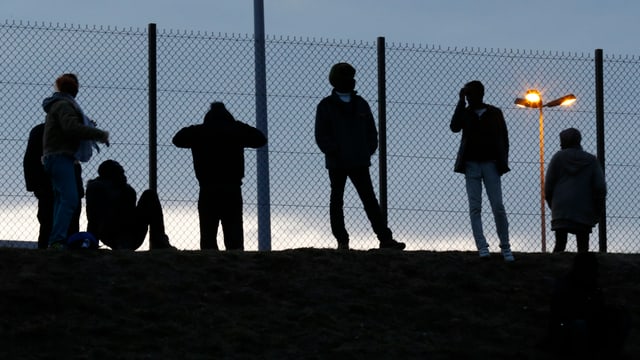 Flüchtlinge stehen an einem Zaun.