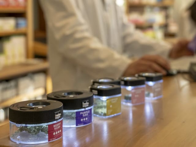 Tresen in einer Apotheke. Verschiedene Gläschen mit Cannabis sind aufgereiht, im Hintergrund steht der Verkäufer.