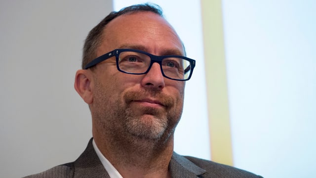 Jimmy Wales mit Anzug und Brille vor einer weissen Wand.