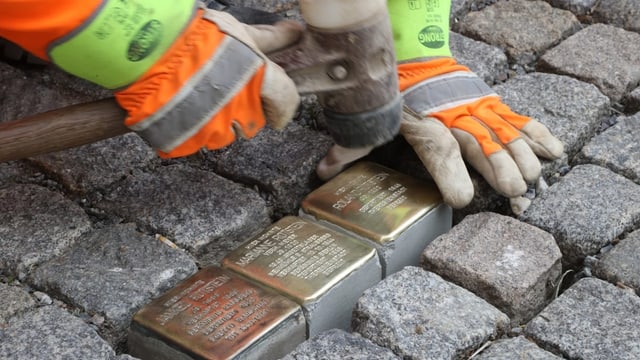 Boden, 3 goldene Steine werden eingelegt. Man sieht Hände in Handschuhen und einen Hammer.