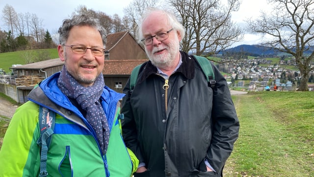 Zwei Männer mittleren Alters in der Natur. Im Hintergrund ist ein Dorf (Herisau) zu sehen. 