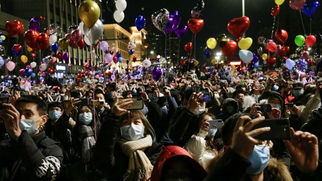 Crowd of people in the dark with balloons.  People wear masks and take selfies.