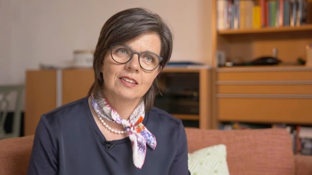 Eine Frau mit Brille und Foulard sitzt auf einem Sofa.