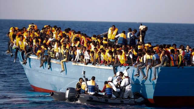 Ein Schlauchboot mit Soldaten vor einem überfüllten Holzboot.