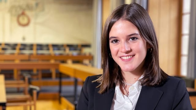 Junge Frau mit dunklen, langen Haaren lächelt in Kamera. Dahinter verschwommen ein Saal mit Sitzbänken.