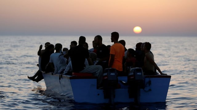 Ein blaues Boot fährt auf weiter See, vollgestopft mit Menschen. Im Horizont geht die Sonne gerade auf.