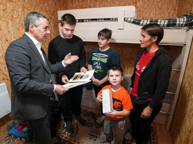 Ignazio Cassis steht in einem Raum mit Holzwänden. Er spricht mit einer Familie.