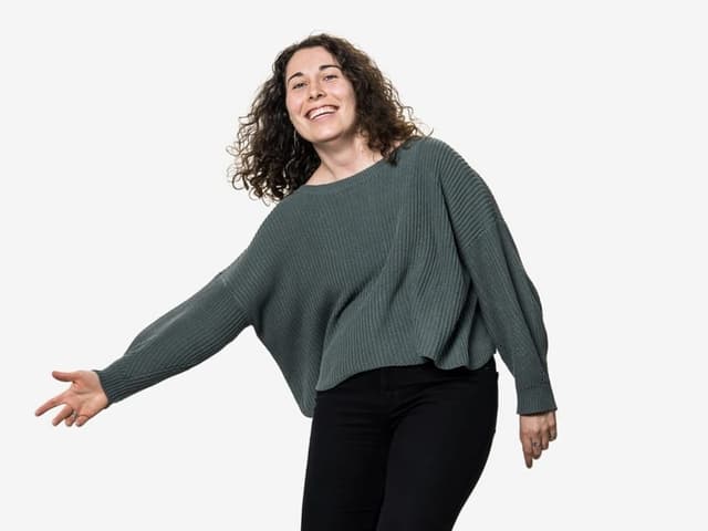 Luana schlägt mit der rechten Hand seitlich in die Luft und schaut in die Kamera. Sie trägt einen grünen Pullover.