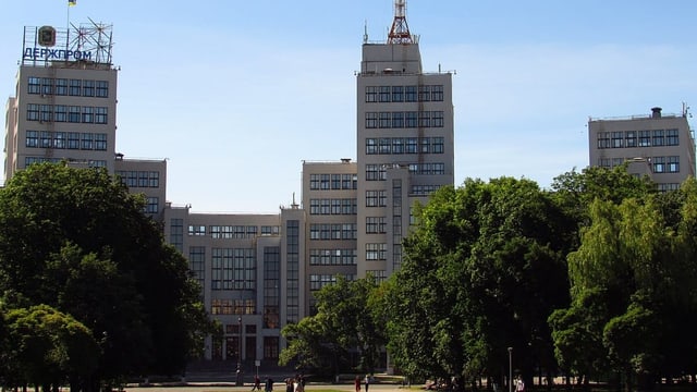 Graue Bettongebäude mit bläulichen Fenstern. Davor grüne Bäume und eine Fussgängerstrasse