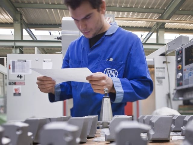 Ein Mann liest in einer Werkstatt einen Zettel. Vor ihm stehen gefräste Teile.