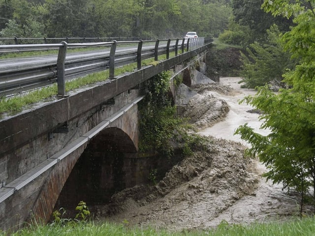 Blick auf eine Brücke, unter der ein Fluss Hochwasser führt. Das braune Wasser staut sich an den Pfeilern.