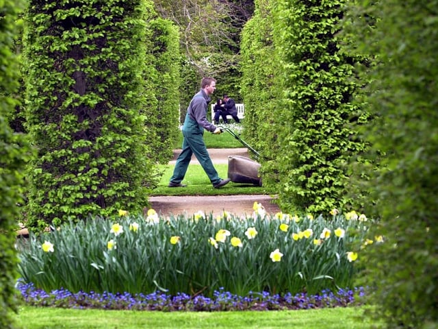 Gärtner läuft im Hintergrund mit Rasenmäher durch Garten.