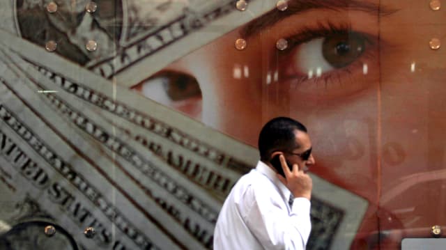 Ein Mann in einem weissen Hemd und mit Sonnenbrille telefoniert vor einem Plakat mit Dollars und Frauenaugen.