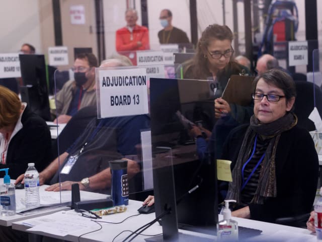 Personen in einem Raum sitzen an Computern hinter dem Schild 'ADJUDICATION BOARD 13'.