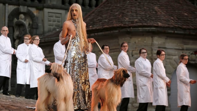 Eine trans Person mit langem Haar und Glitzerkleid führt zwei Hunde Gassi. Dahinter stehen Mediziner in Arztkittel. 