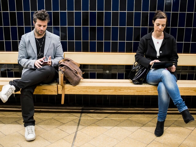 Ein Mann und eine Frau sitzen auf einer Bank. Sie schauen beide auf ein Smartphone bzw Tablet. 