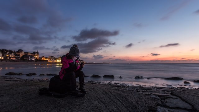 Frau sitzt in der Dämmerung auf einem Stein am Strand und schaut aufs Smartphone.