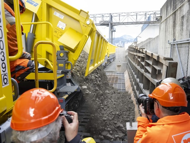 Arbeiter in Schutzhelmen beobachten Steine, die aus gelbem Fahrzeug entladen werden.