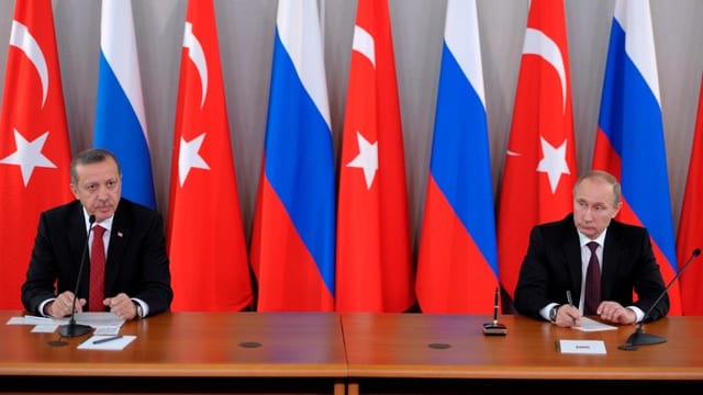 Erdogan und Putin sitzen an einem Tisch, offensichtlich anlässlich einer Medienkonferenz, hinter ihnen russische und türkische Fahnen.