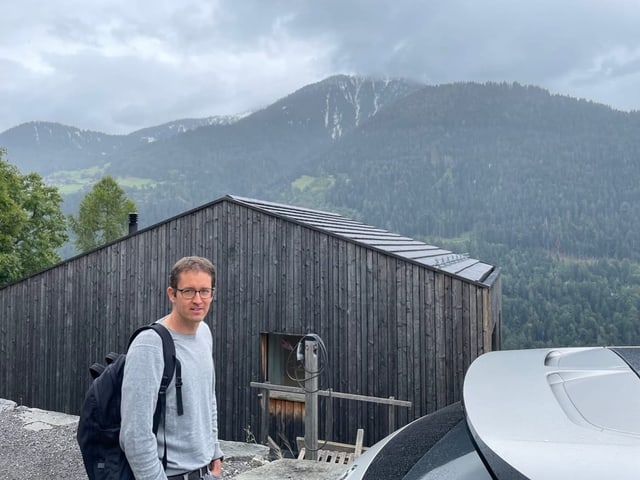 Eine Person und ein Auto vor einem Haus mit Bergen im Hintergrund.