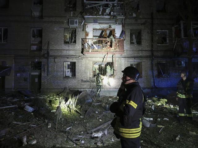 Feuerwehrmänner vor beschädigtem Gebäude bei Nacht.