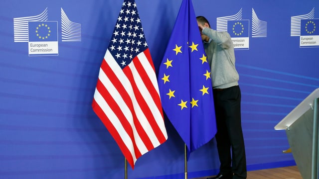 Die Amerikanische und die EU-Flagge nebeneinander. Die EU-Flagge wird von einem Mann gerichtet.