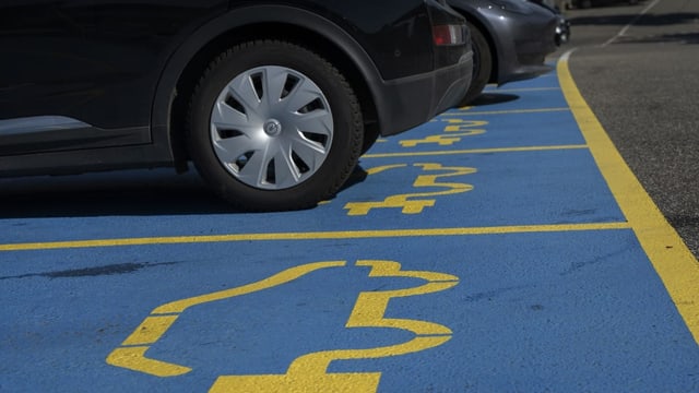 Viele Autos sind von unten zu sehen, die auf blauen Parkfeldern stehen. Ein gelbes Symbol darauf zeigt ein E-Auto an.