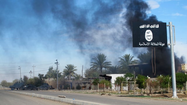 Zu sehen ist eine schwarze IS-Flagge mit weissen arabischen Schriftzeichen. Dahinter eine Stadt mit Palmen, aus welcher schwarzer Rauch aufsteigt