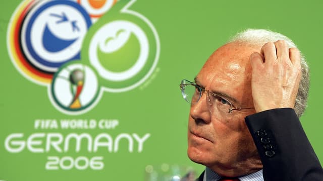 Beckenbauer kratzt sich am Kopf, hinter ihm das Logo der Fussball-WM 2006.