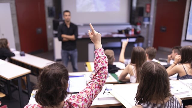 In einem Klassenzimmer steht ein Lehrer, auf Schülerinnen und Schüler sitzen in den Reihen, zwei melden sich.